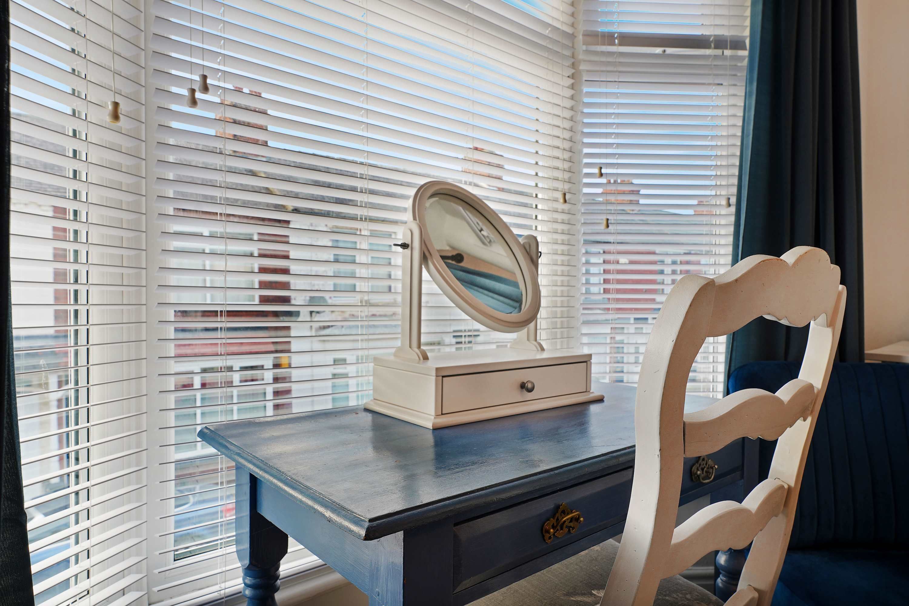 Ein Zimmer mit Fenster, Jalousien, einem Schminktisch mit Spiegel und einem Stuhl.
