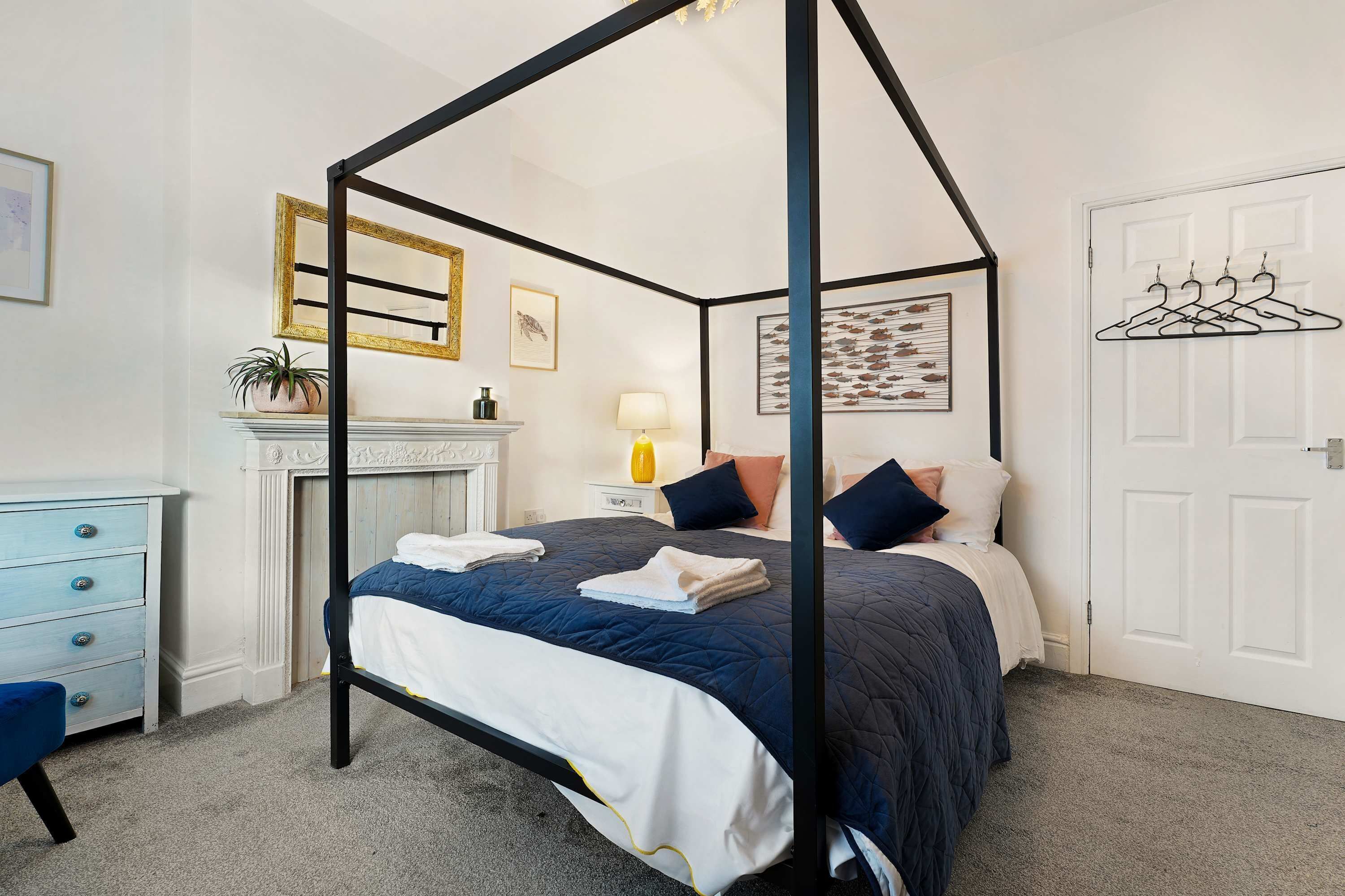 A bedroom with a four-poster bed, a dresser, a mirror, a fireplace, and a door with a coat rack.