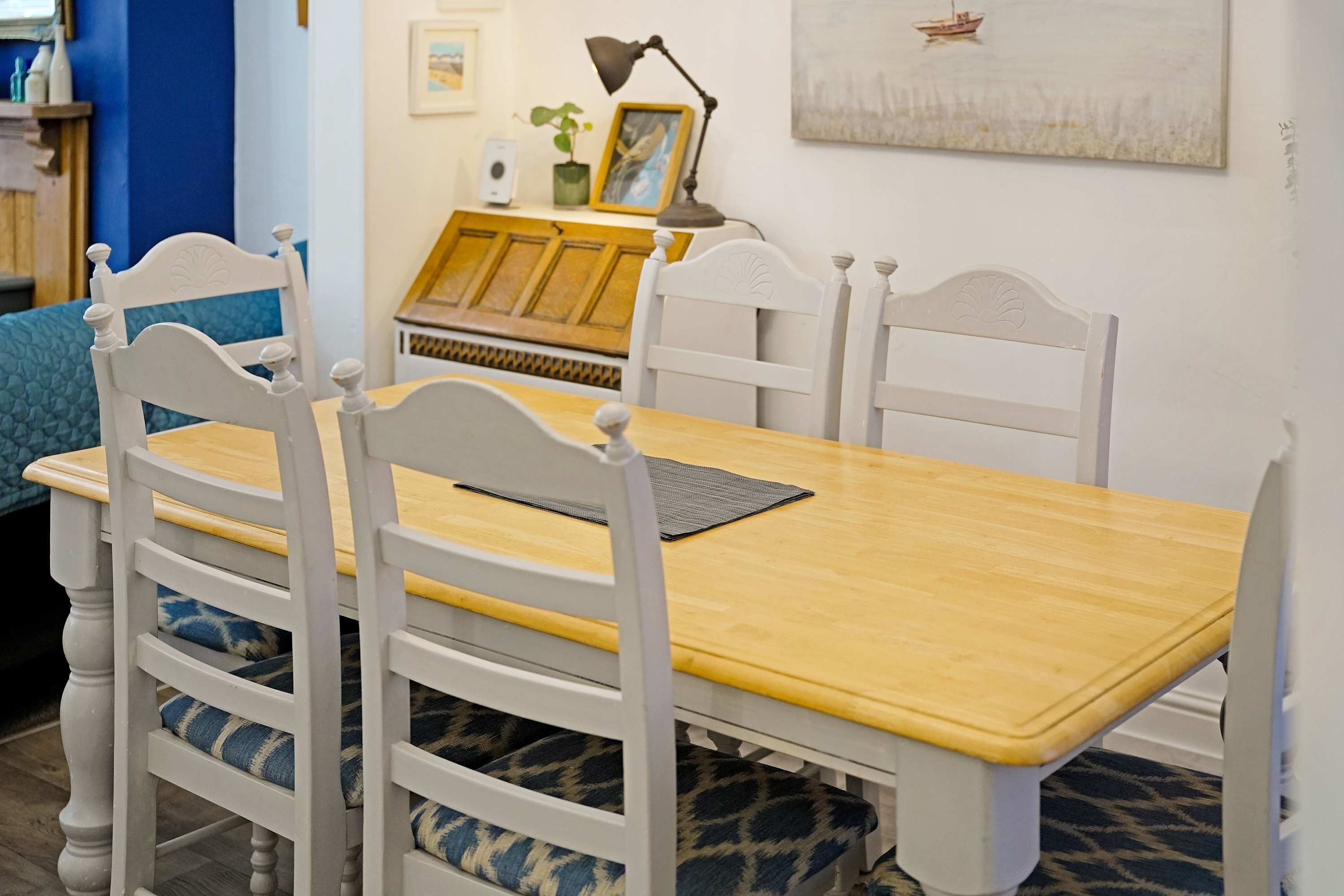 A dining table with four white chairs, a yellow tablecloth, and a framed picture on the wall.