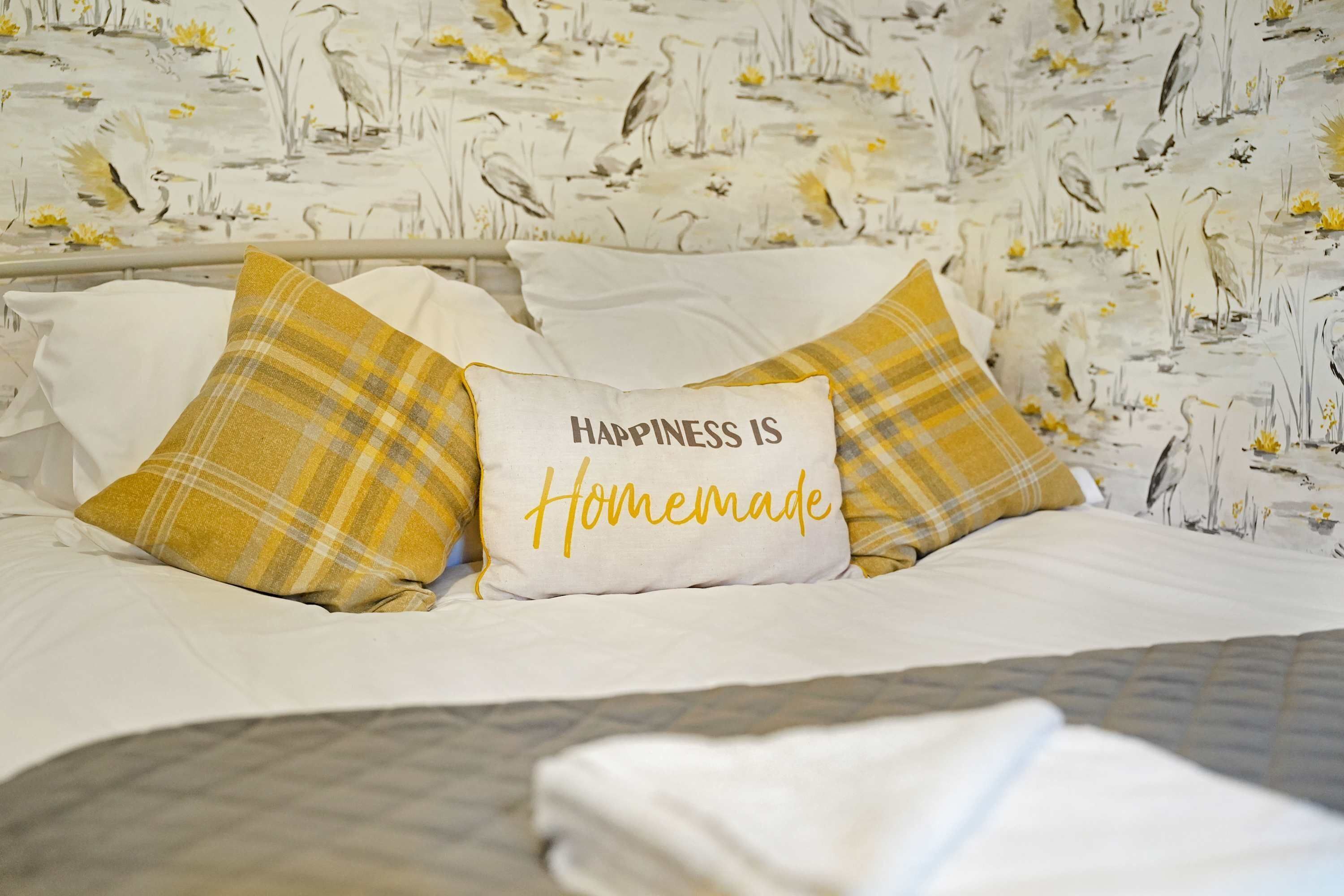 A bed with white bedding and two yellow plaid pillows, one with the text 'Happiness is Homemade'.
