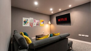 A modern living room with a grey sofa, colorful cushions, a wall-mounted TV displaying 'NETFLIX', and a series of framed artworks on the wall.