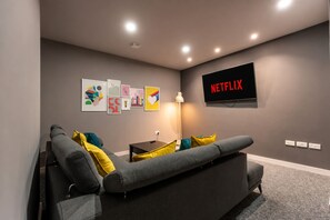 A modern living room with a grey sofa, colorful cushions, a wall-mounted TV displaying 'NETFLIX', and a series of framed artworks on the wall.