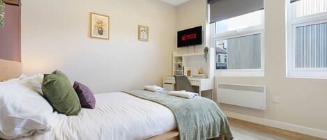 A bedroom with a bed, a TV, a window with blinds, and a radiator.