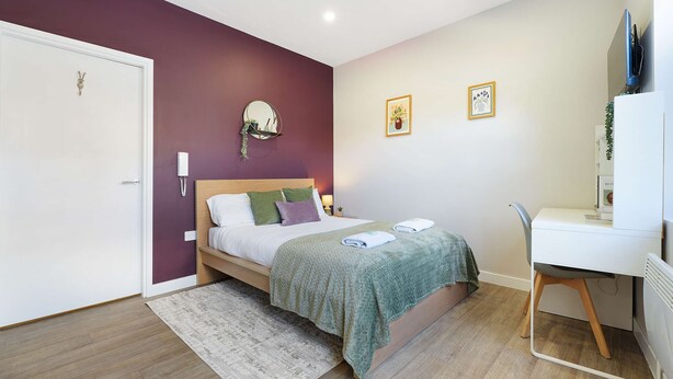 A bedroom with a bed, bedside table, chair, desk, and a mirror.