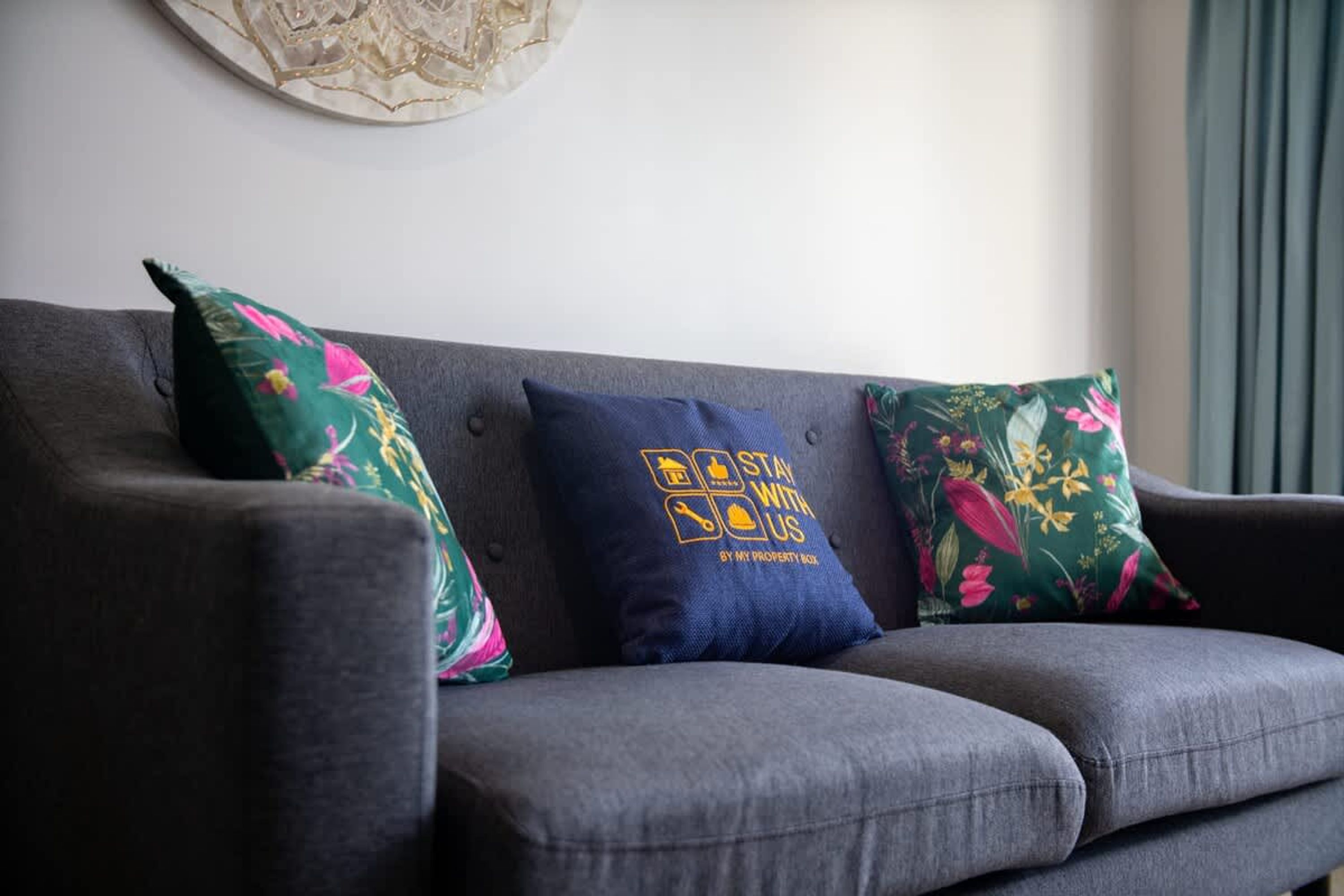 A grey sofa with three cushions, one with a floral pattern and another with a blue cushion featuring a logo and text.