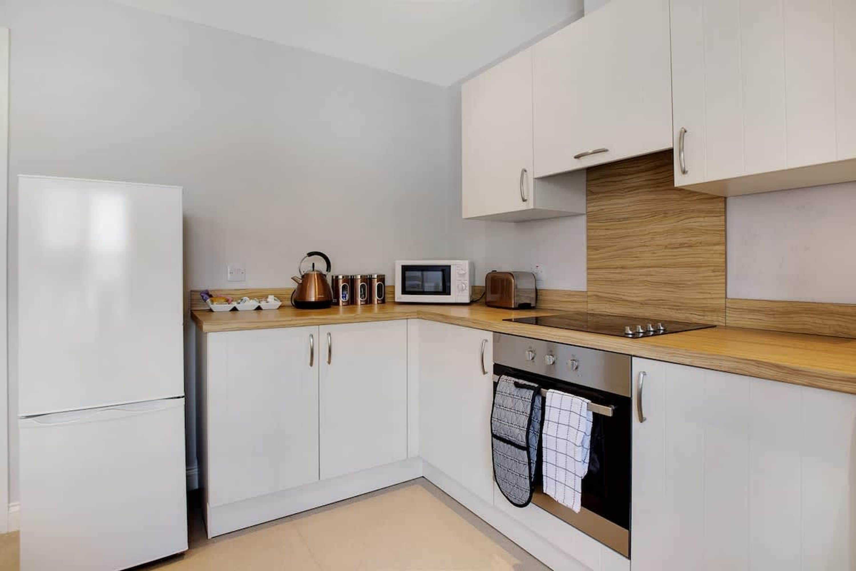 Una cocina moderna con armarios blancos, encimera de madera, refrigerador de acero inoxidable, microondas, hervidor y horno empotrado.