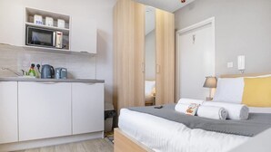 Habitación de hotel compacta con kitchenette, cama con almohadas y manta, y mesita de noche con lámpara.