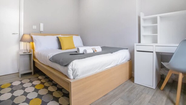 A bedroom with a bed, bedside table, lamp, and a desk with a chair.