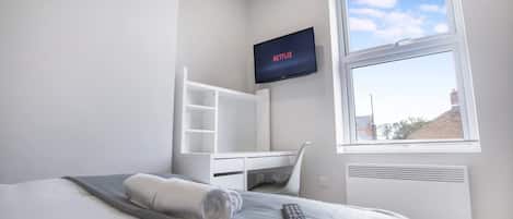 A hotel room with a bed, a TV mounted on the wall, a window with a view, and a white desk with shelves.