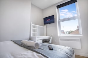 A hotel room with a bed, a TV mounted on the wall, a window with a view, and a white desk with shelves.