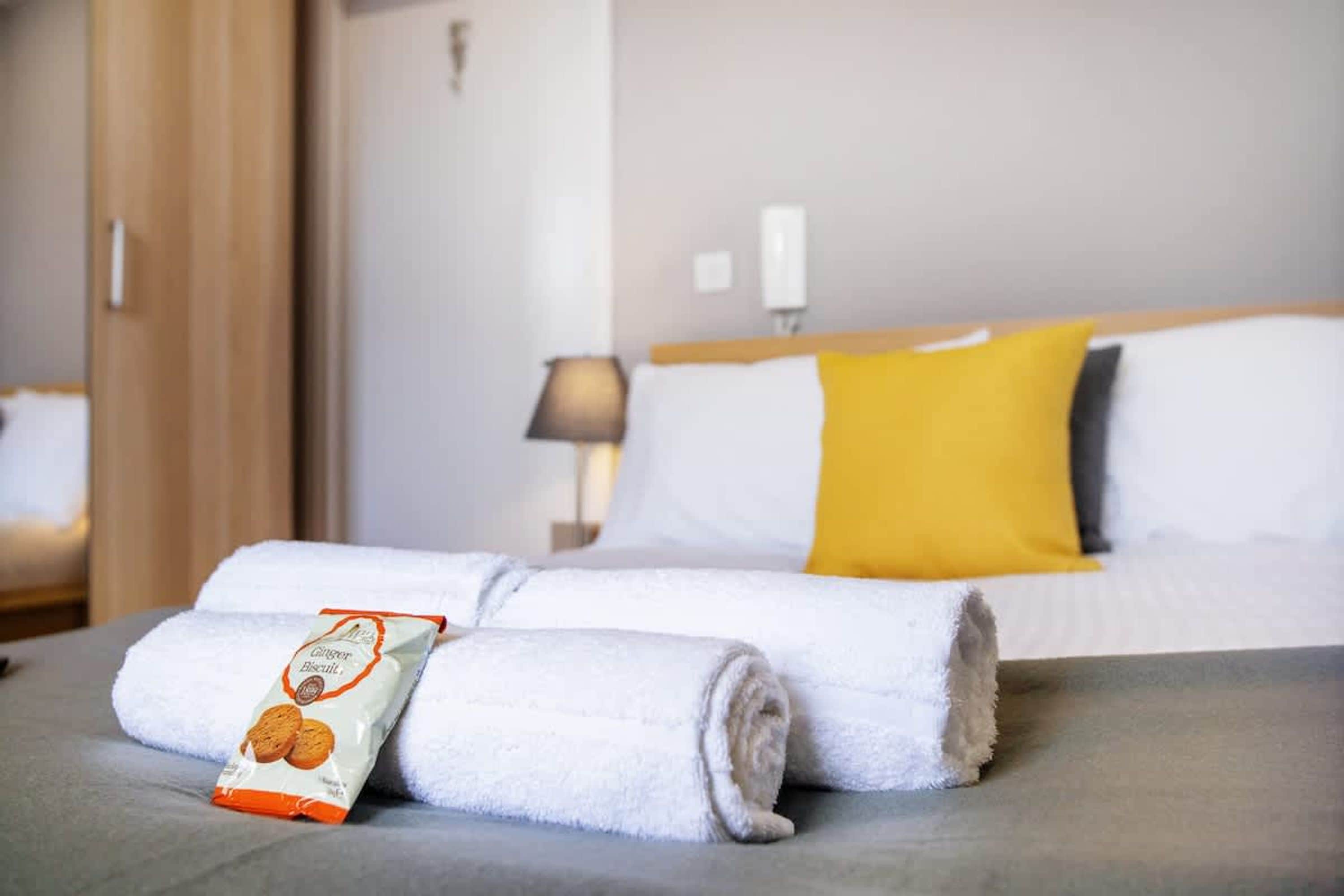 A neatly made bed with a yellow pillow, a white pillow, and a white blanket rolled up at the foot. A package of ginger biscuits is placed on the bed.