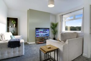 Un salon avec un canapé, une petite table, un téléviseur posé sur un meuble, une plante en pot et une fenêtre donnant sur un beau paysage.