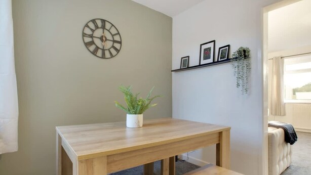 A room with a wooden table and bench, a wall-mounted clock, a shelf with framed pictures, and a hanging plant. - Family two Bedroom Home Free Parking and Backyard (Crook)