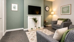 A living room with a fireplace, a television mounted above it, a clock on the wall, and a sofa. - Modern 2 Bed Near Seafront and Easy Commute Links (South Shields)