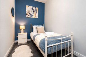 A bedroom with a white bed, a bedside table with a lamp, a blue wall, and a painting. - Modern 2 Bed Near Seafront and Easy Commute Links (South Shields)