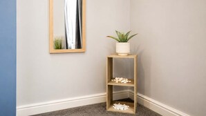 A corner with a wooden floor, a white wall, a mirror with a wooden frame, a small wooden shelf with a plant and decorative items, and a dark carpet. - Modern 2 Bed Near Seafront and Easy Commute Links (South Shields)