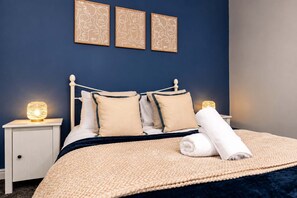 A neatly made bed with a dark blue headboard, two bedside tables with lamps, and three framed wall art pieces above the bed. - Modern 2 Bed Near Seafront and Easy Commute Links (South Shields)
