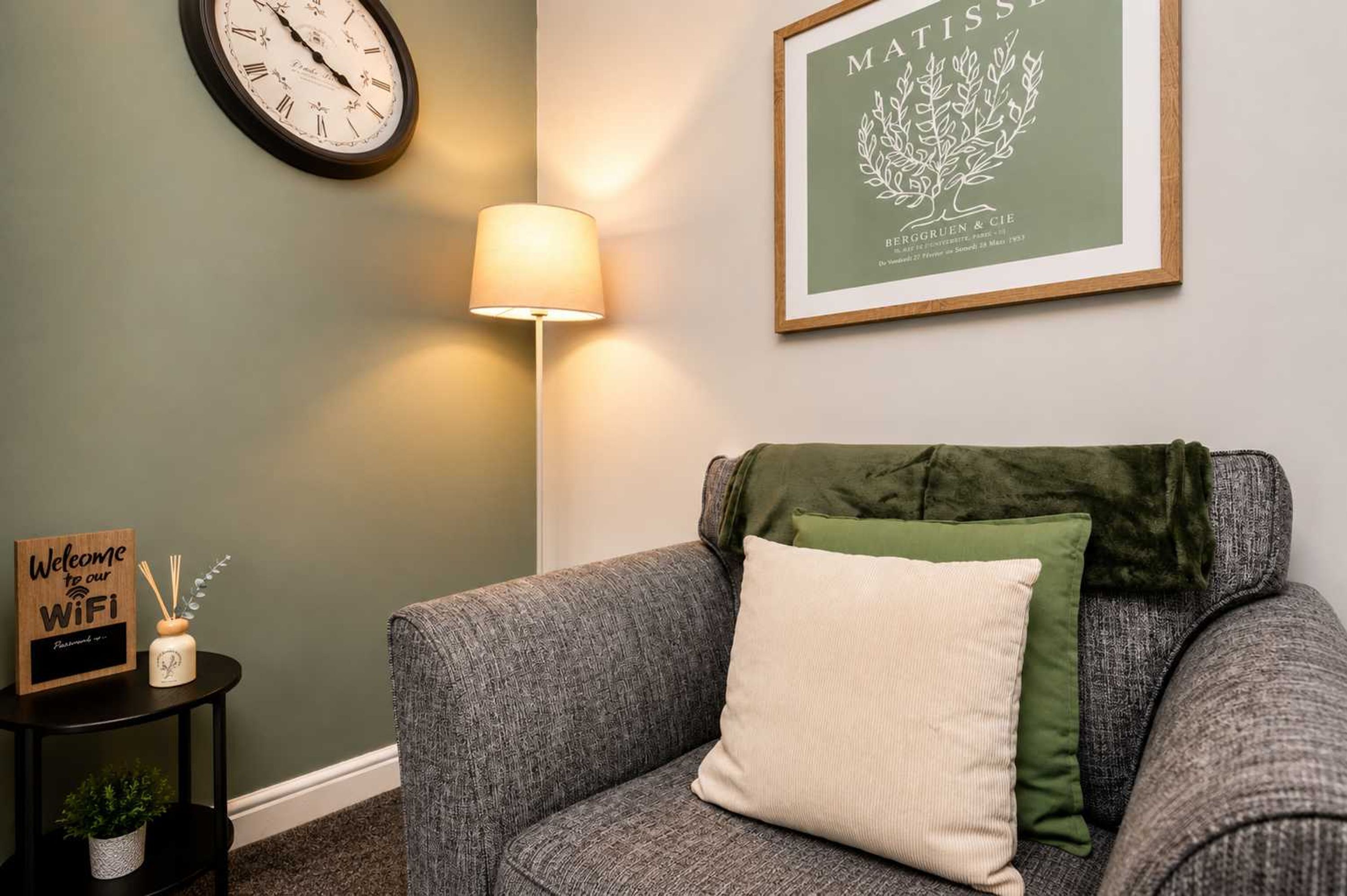 A room with a grey sofa, a wall clock, a framed artwork, a lamp, and a sign indicating WiFi availability.