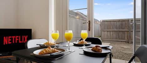 Un comedor con una mesa dispuesta para el desayuno, que incluye croissants y vasos de zumo de naranja, y un televisor mostrando el logotipo de Netflix.