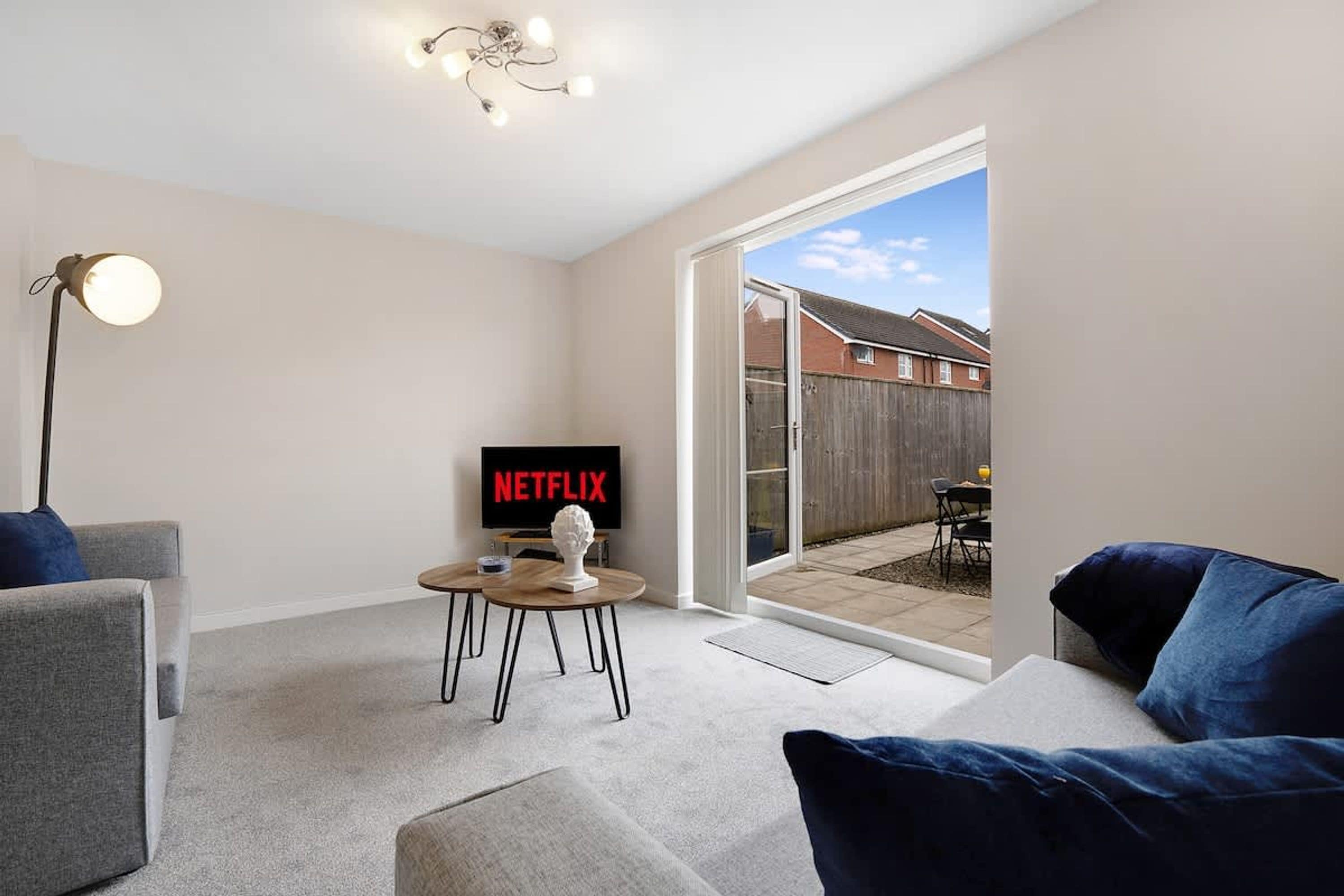 Een woonkamer met een flatscreen-tv waarop 'NETFLIX' in rode letters staat, een vloerlamp en een klein rond tafeltje met een witte sculptuur.