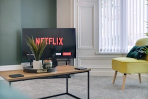 Ein Wohnzimmer mit Fernseher, der das Netflix-Logo zeigt, einem kleinen Tisch mit einer Pflanze, einem gelben Sessel und einem Fenster mit Jalousien.