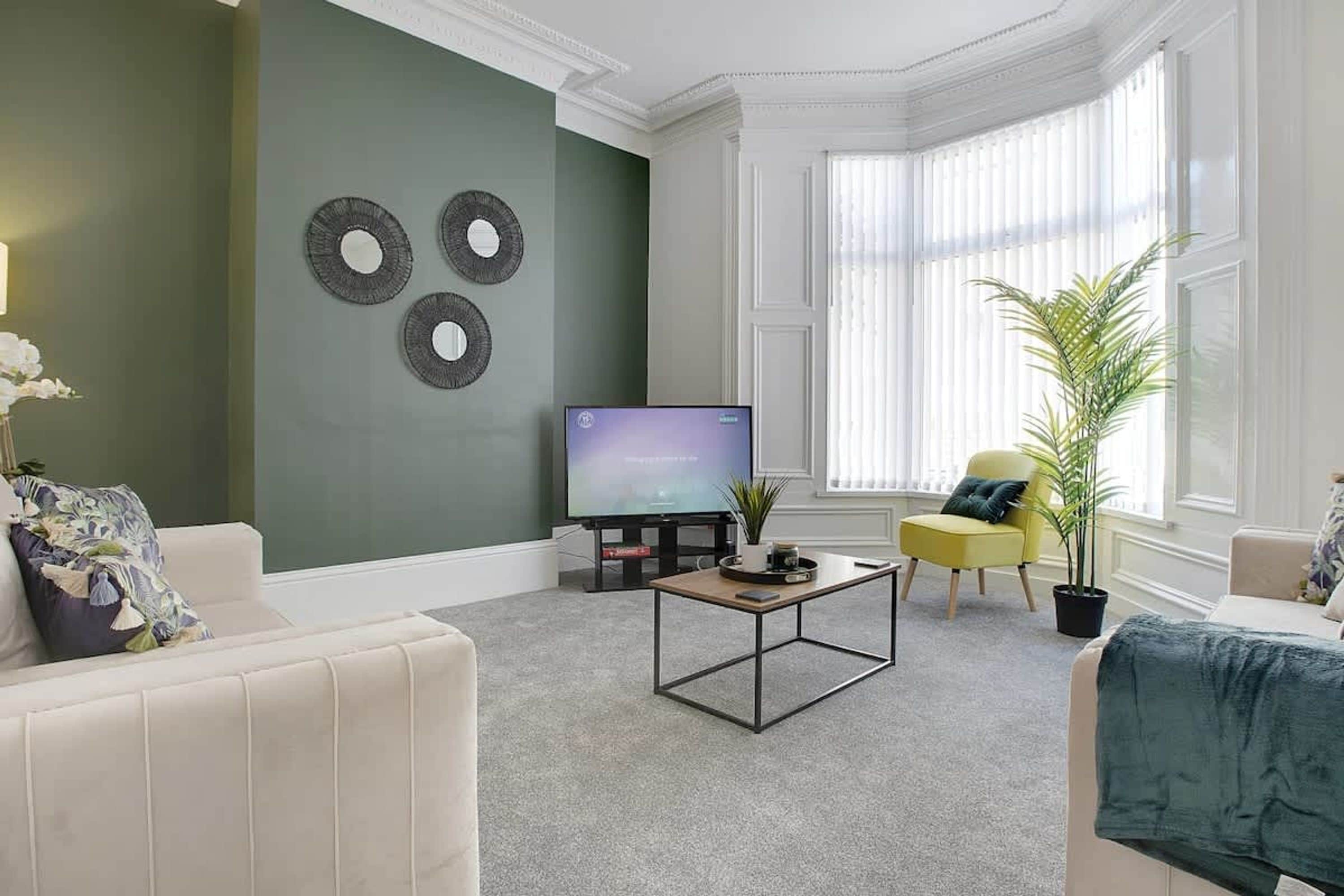 A living room with a flat-screen TV, a coffee table, a sofa, a yellow armchair, and a large window with blinds.