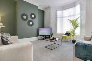 Un salón con televisión de pantalla plana, una mesa de centro, un sofá, un sillón amarillo y un ventanal con persianas.