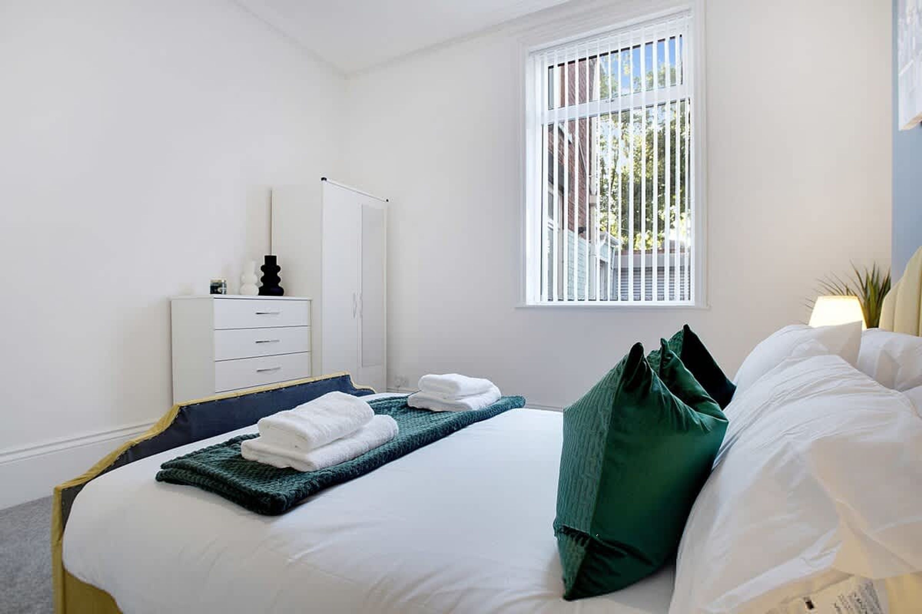 Une chambre d’hôtel avec un lit, une commode, une fenêtre avec des stores et une lampe.