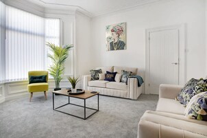 Una sala de estar con un sillón amarillo, un sofá blanco, una mesa de centro y un ventanal con persianas.