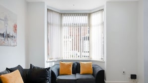 A modern living room with a corner window, a black sofa with two cushions, and a wooden coffee table. - Classic 4 Bedroom Retreat Close to Infinity Bridge (Stockton-on-Tees)
