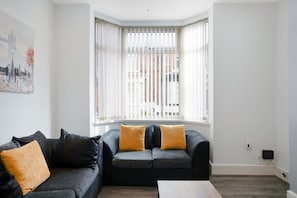 A modern living room with a corner window, a black sofa with two cushions, and a wooden coffee table. - Classic 4 Bedroom Retreat Close to Infinity Bridge (Stockton-on-Tees)