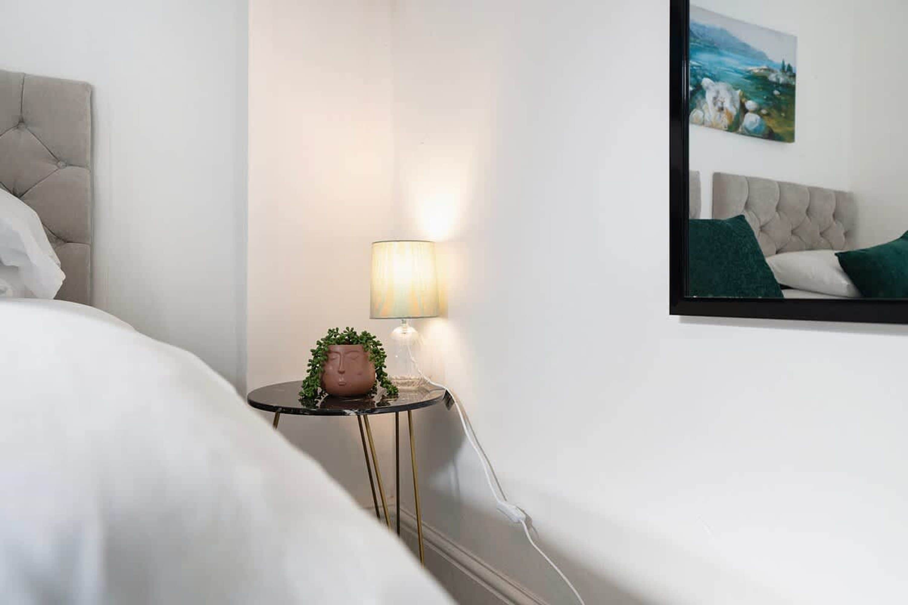 A hotel room with a bed, a bedside table with a lamp, a small potted plant, and a painting on the wall.