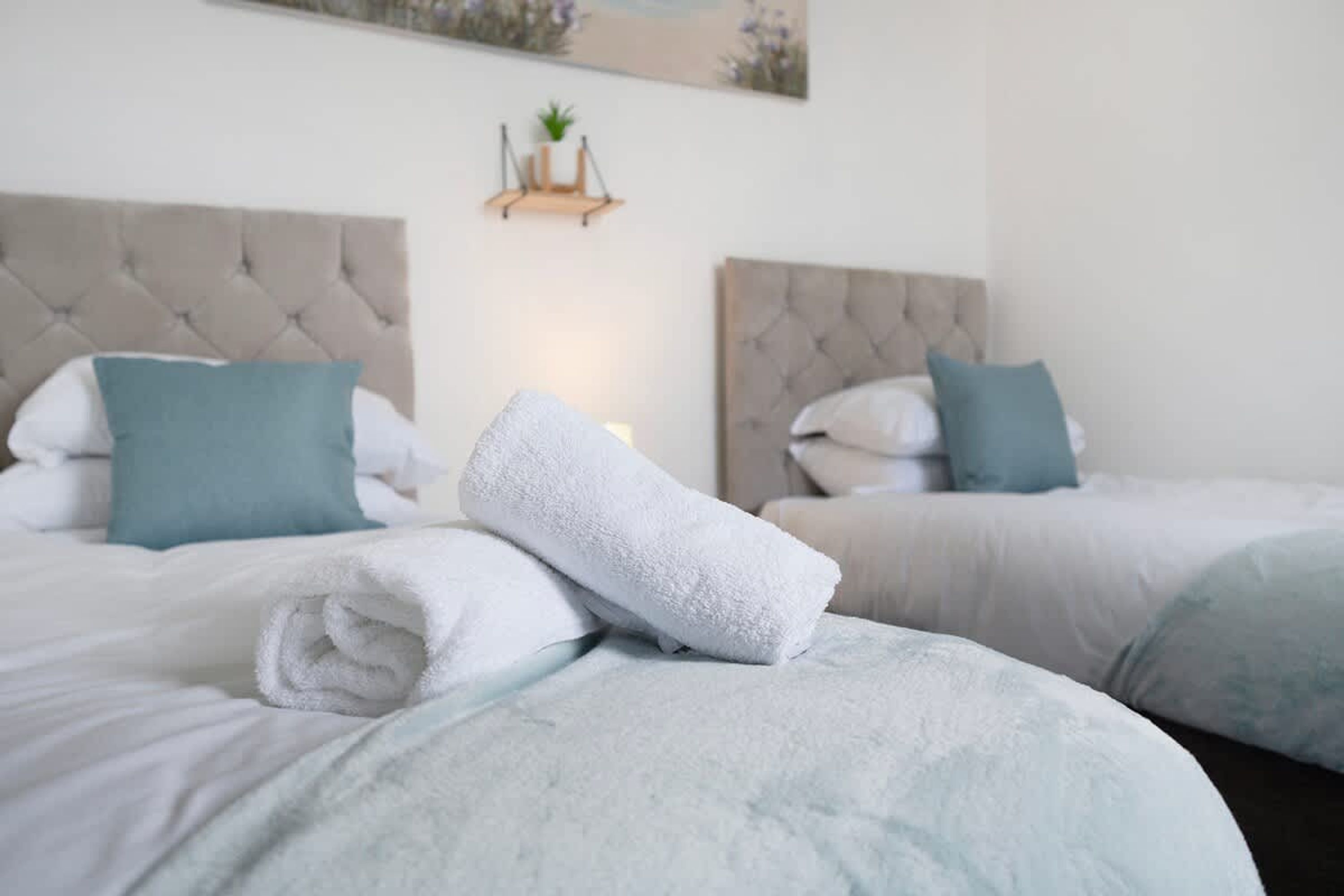 A hotel room with two beds, each with a tufted headboard, white bedding, and teal pillows. A rolled white towel is placed on the bed in the foreground.