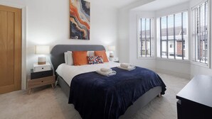 A modern bedroom with a large bed, two bedside tables, a nightstand, a painting, and a view of the outside through large windows. - Central Newcastle 2BR With Free Parking and Wifi (Newcastle-upon-Tyne)