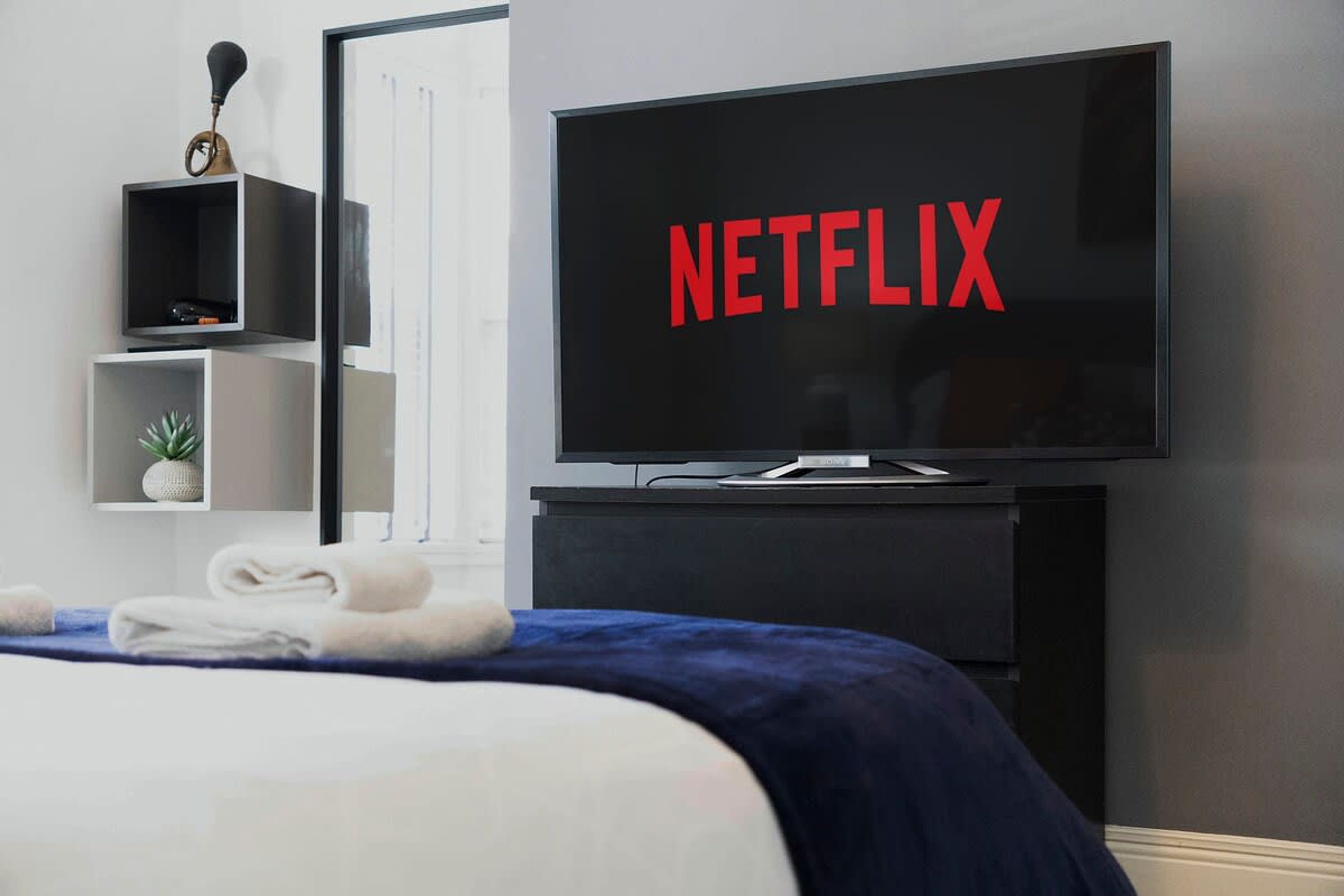 Une chambre d’hôtel avec un lit, un téléviseur à écran plat fixé sur un support noir et un mur orné d’une étagère sur laquelle se trouve une plante.