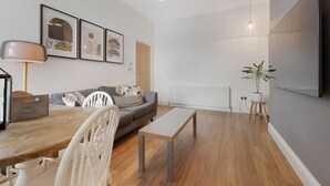 A modern living room with a sofa, a dining table, and a coffee table. There are framed pictures on the wall, a lamp, and a potted plant. - Central Newcastle 2BR With Free Parking and Wifi (Newcastle-upon-Tyne)