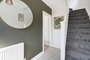 Un couloir avec un mur vert foncé, une porte blanche, un escalier recouvert d’une moquette sombre et un miroir rond doté d’un cadre doré.