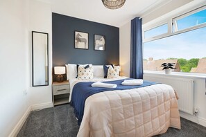 A bedroom with a bed, a bedside table, a lamp, a window with curtains, and a view of the outside. - Stylish Hartlepool 3BR Sleeps 5 by Stay With Us (Stockton-on-Tees)