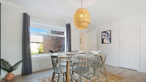 A dining area with a round table, chairs, a woven pendant light, and a view of a brick building through a window. - Stylish Hartlepool 3BR Sleeps 5 by Stay With Us (Stockton-on-Tees)