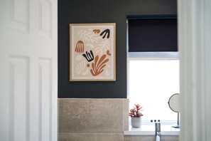 Ein Badezimmer mit einem gerahmten abstrakten Kunstwerk an der Wand, einem Fenster mit Blick und einem Waschbecken mit Spiegel.