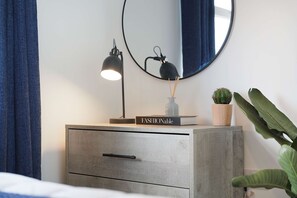 Una mesita de noche con una lámpara, un libro titulado 'FASHIONable', un pequeño cactus en maceta y un espejo en la pared.