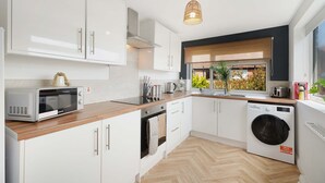 A modern kitchen with white cabinetry, a built-in oven, a microwave, a washing machine, and a window with a view of greenery. - Stylish Hartlepool 3BR Sleeps 5 by Stay With Us (Stockton-on-Tees)
