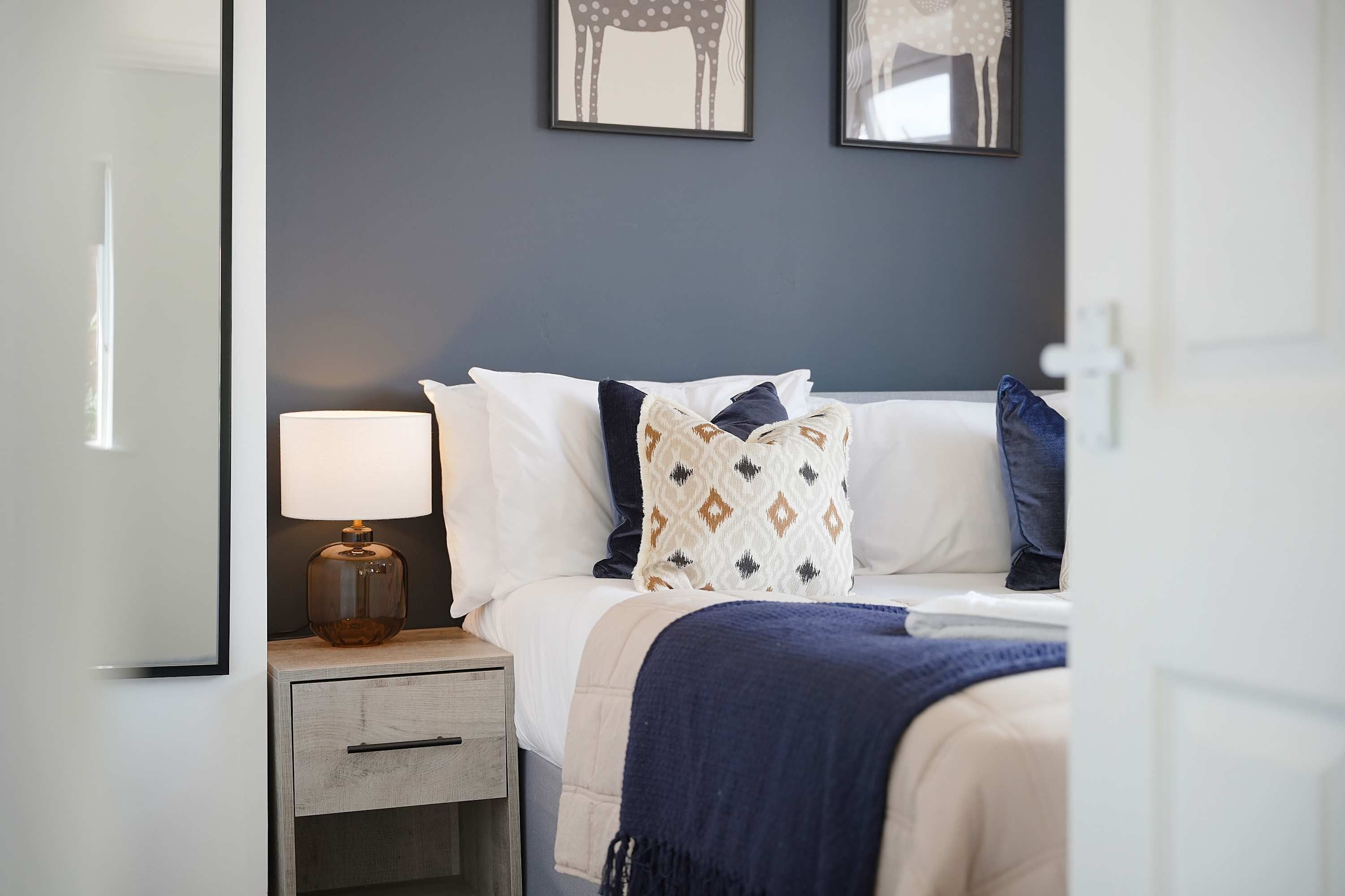 A bedroom with a bed, a bedside table with a lamp, a nightstand, and two wall-mounted mirrors.