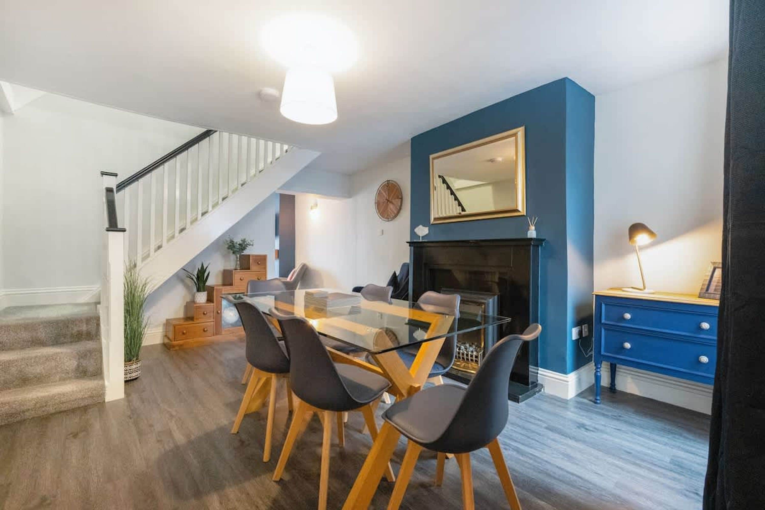 A dining area with a glass table and chairs, a staircase, a blue accent wall, and a fireplace.