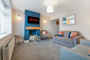 Una sala de estar moderna con una pared azul destacada, un televisor de pantalla plana montado sobre la chimenea, un sofá gris con cojines coloridos, un sillón pequeño y un reloj redondo en la pared.