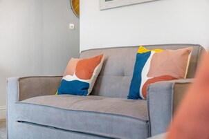 Un sofá gris con dos almohadas coloridas, un cuadro enmarcado en la pared y un interruptor de luz.