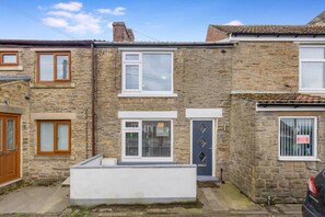 A two-story stone house with a small front porch, a garage door, and a car parked in the driveway. - Stylish 2 Bedroom in Bishop Auckland With Parking (Bishop Auckland)