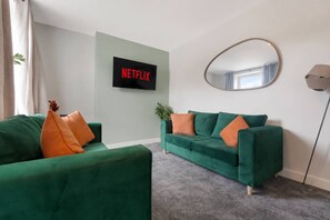 Ein modernes Wohnzimmer mit grünen Sofas, einem Flachbildfernseher, der 'NETFLIX' zeigt, einem runden Spiegel und einer Topfpflanze.