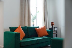 Un divano verde con cuscini arancioni, una pianta in vaso e un vaso con fiori vicino a una finestra.