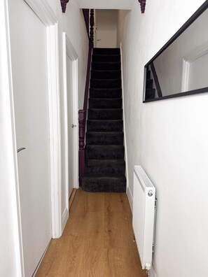 Ein enger Flur mit Holzboden, einem Teppich aus schwarzem Flor und einer schwarzen Treppe, sowie weißen Wänden und einem weißen Heizkörper.